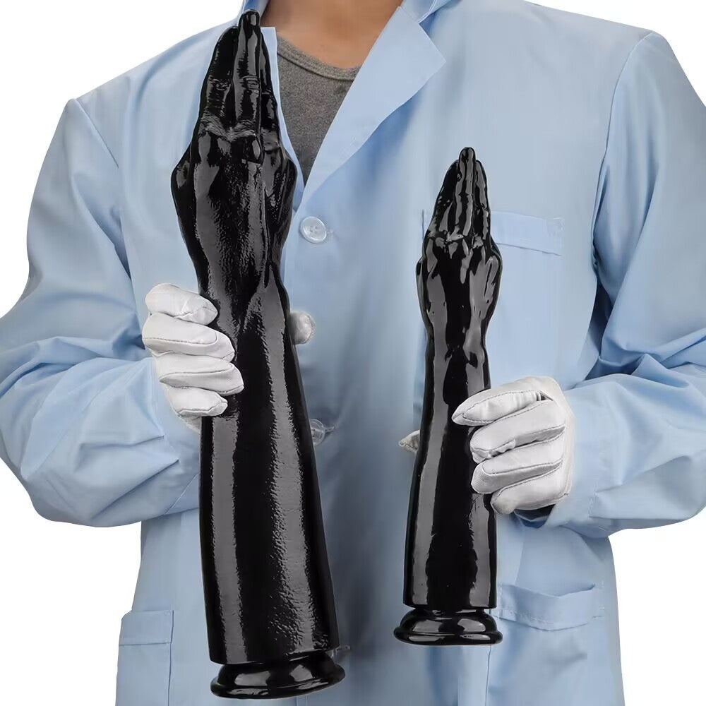 A person wearing a lab coat and white gloves holding the large and small versions of the black prayer hand arm dildo for size comparison.