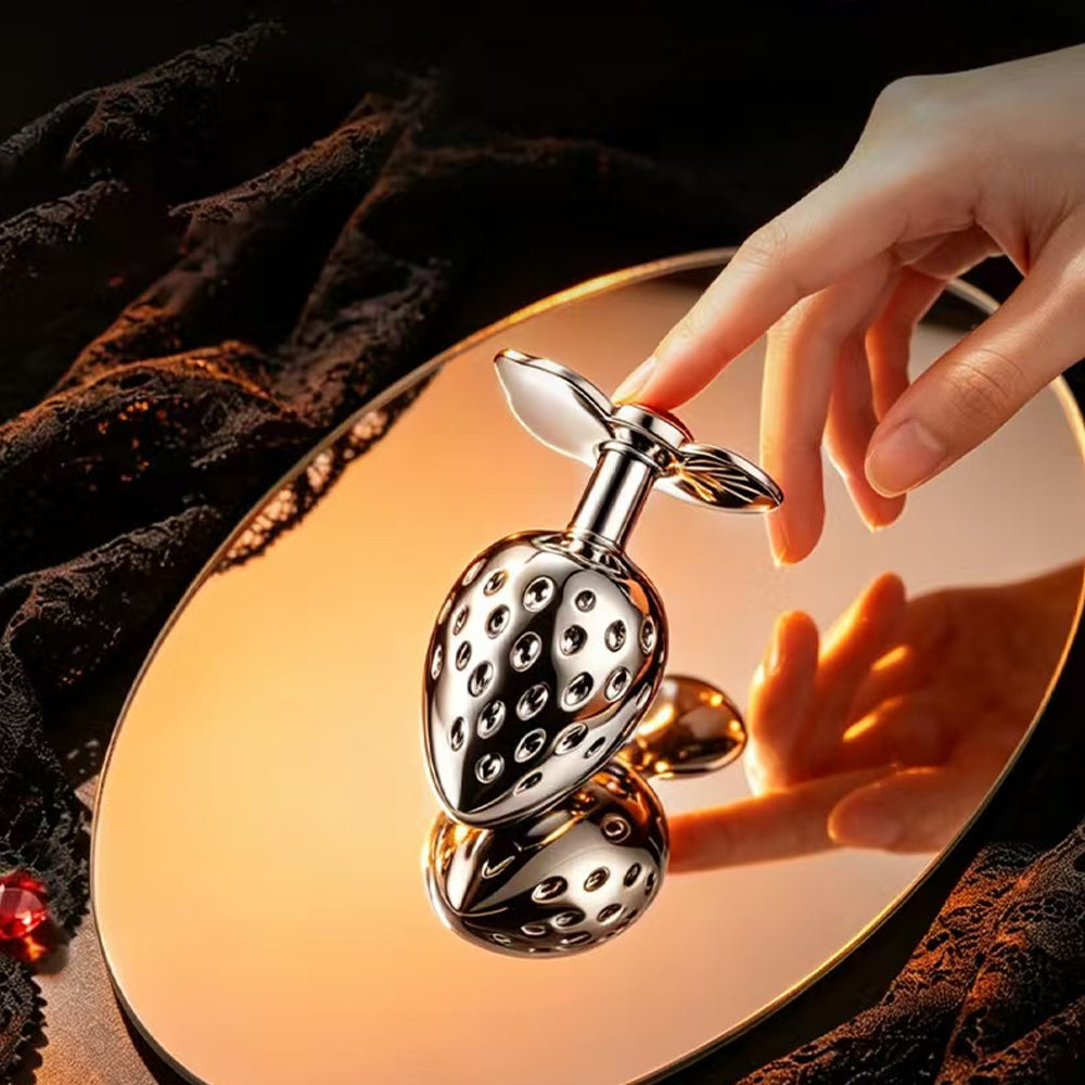 A hand touching a luxury strawberry metal butt plug resting on a gold mirror, showing its flawless reflective surface and premium jewelry-like design.