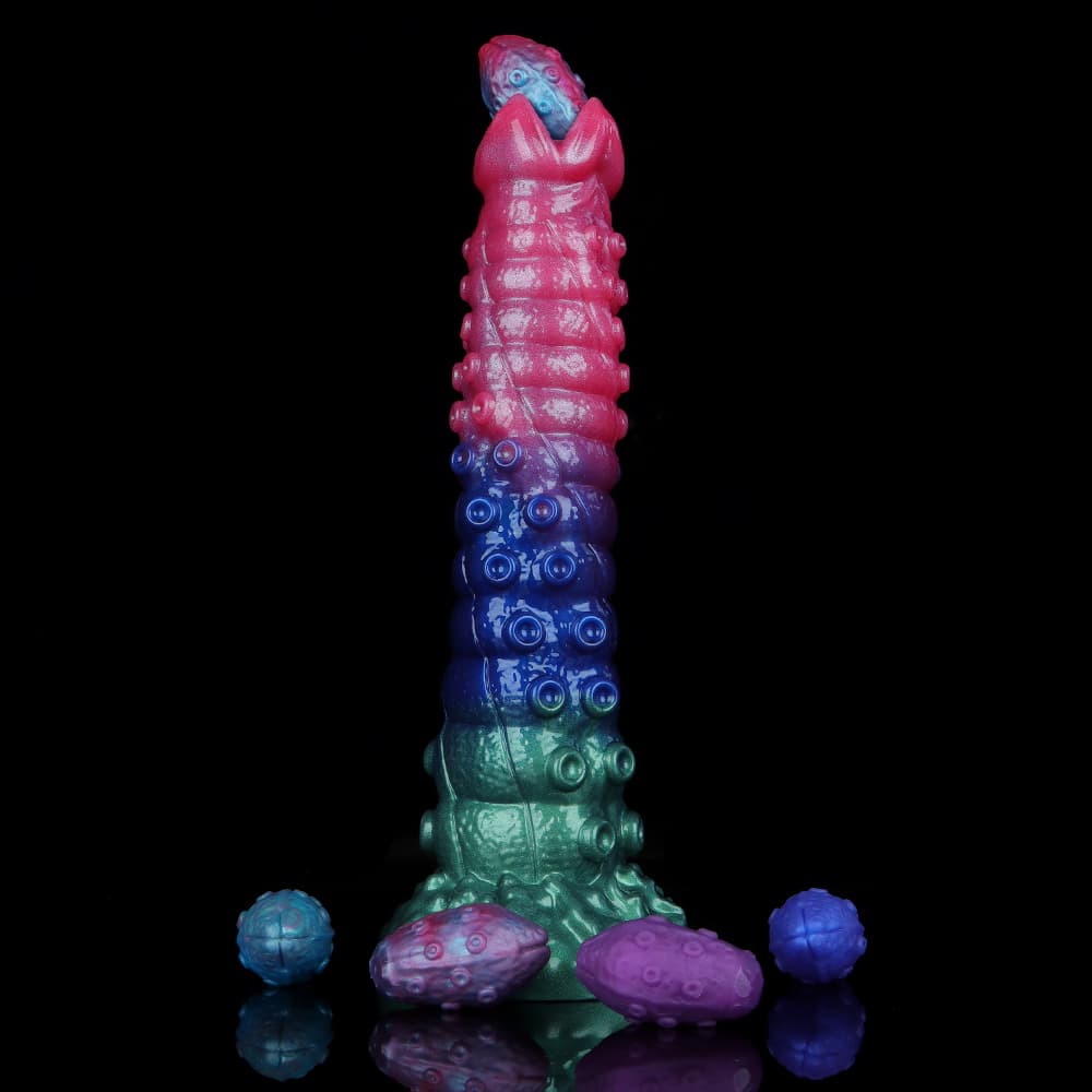 Standing tri-color alien egg laying dildo with a blue silicone egg emerging from the claw tip, surrounded by purple and pink eggs on a black surface.