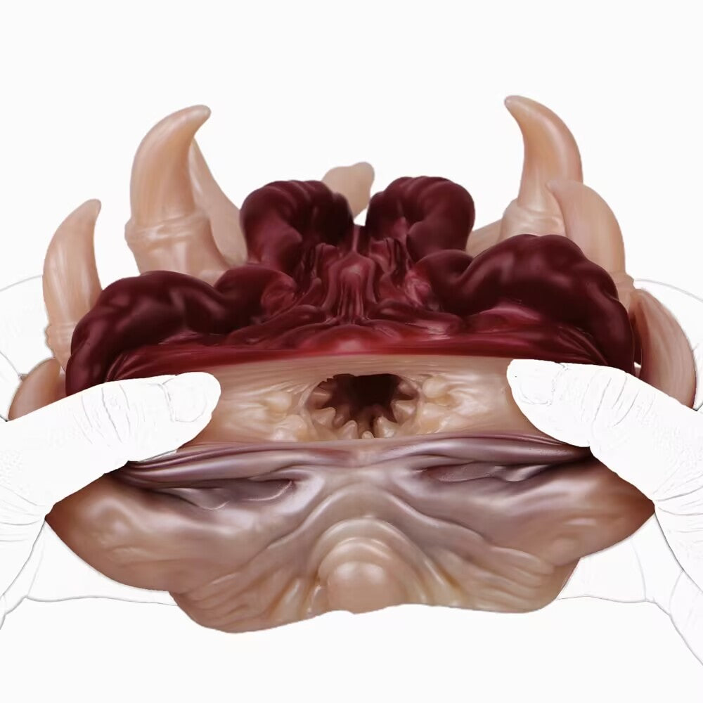 A close-up view of the Alien Facehugger-themed silicone stroker toy, with hands pulling the edges of the opening wide to demonstrate the elasticity. The deep red inner texture is prominent.