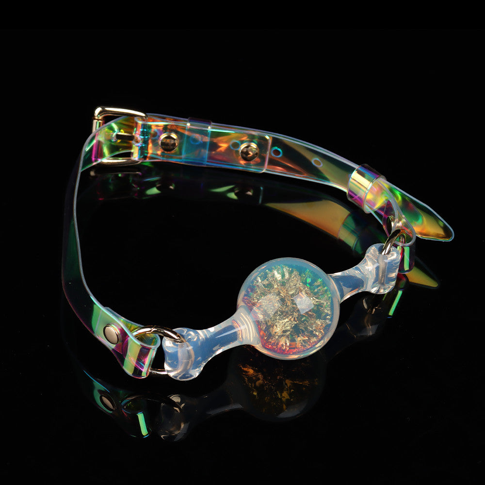 Close-up shot of a clear silicone mouth gag filled with gold glitter foil flakes, featuring an adjustable holographic strap with silver rivets.