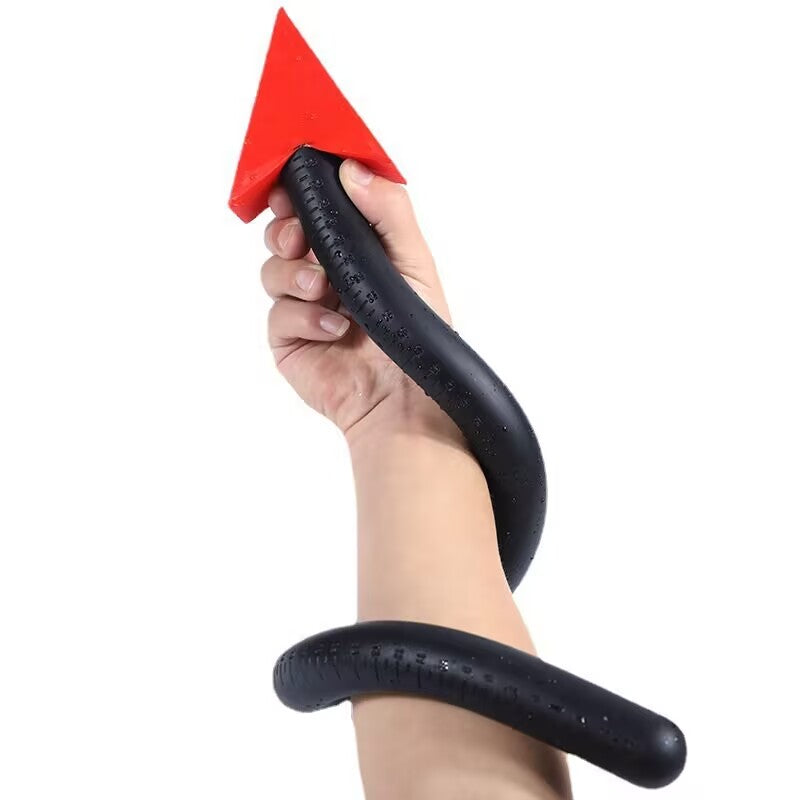 A highly flexible black silicone devil tail anal probe, featuring centimeter depth scale markings, is bent into an S-curve and wrapped around a person's wrist against a white background. The vibrant red arrowhead base acts as a stopper. The soft TPE/silicone material looks supple.