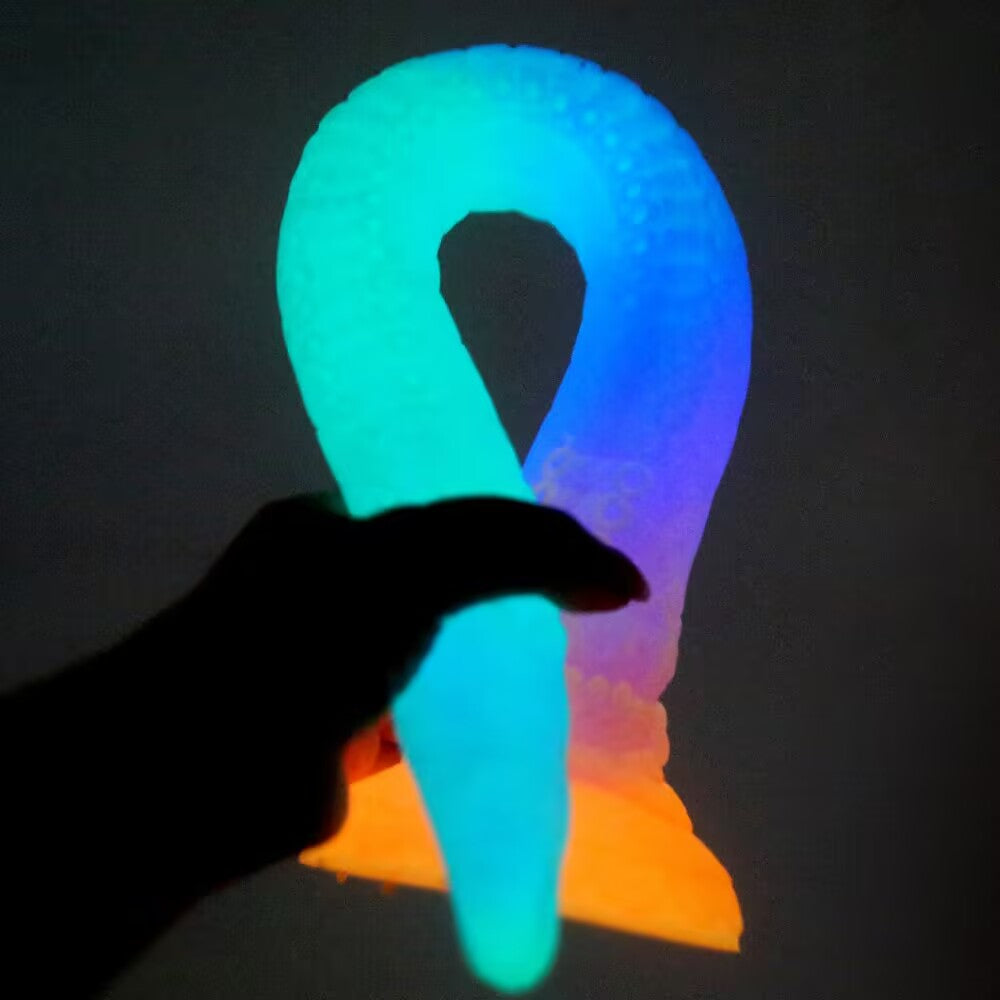 A hand holding a glowing neon blue, purple, and orange fluorescent monster tentacle dildo in a dark room, highlighting its glow-in-the-dark effect.