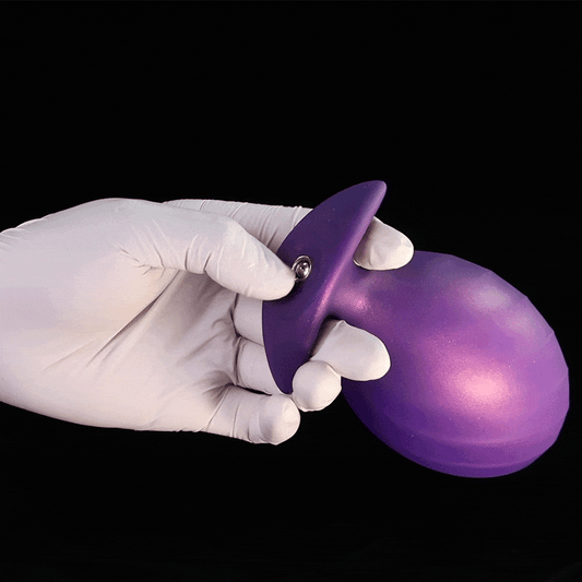 A hand wearing a white glove holds a purple inflatable silicone butt plug, showing the close-up details of the secure metal air valve connector on the anchor base.