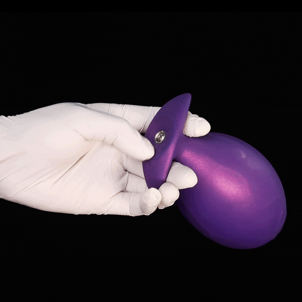 A gloved hand holding a deflated purple inflatable fantasy butt plug showing its slim starting shape and metal air valve before expansion.