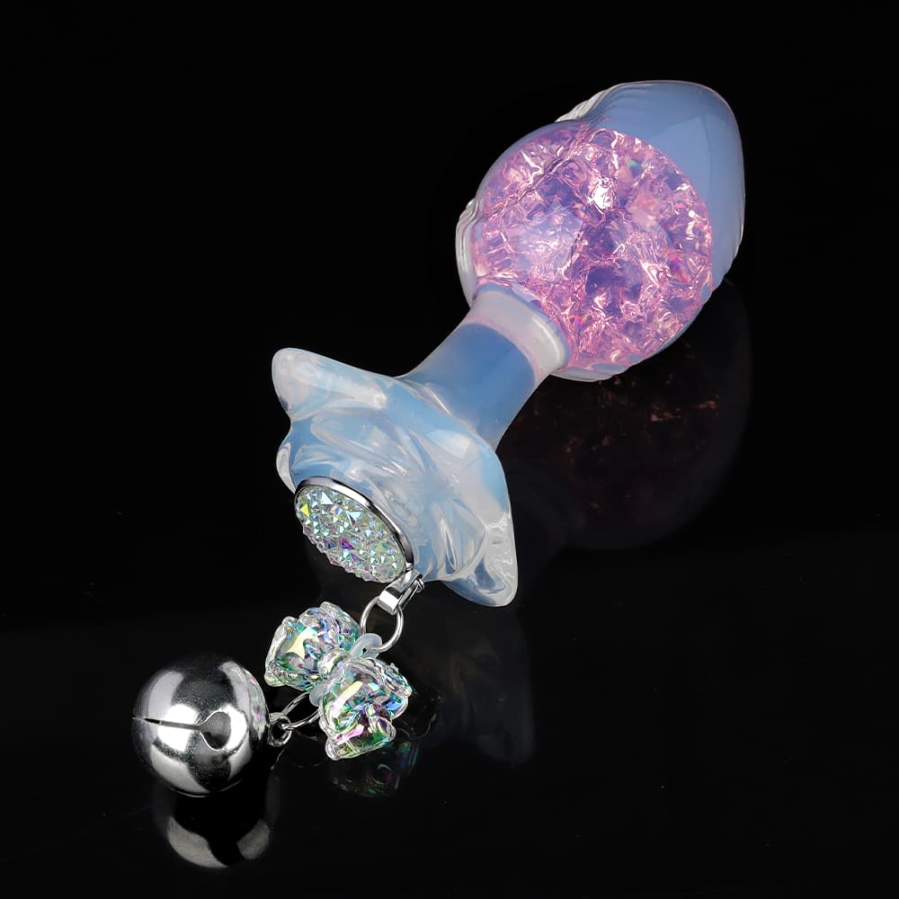 A smooth glass butt plug displaying a glowing pink inner crystal core and a rhinestone rosette base with a ribbon charm.