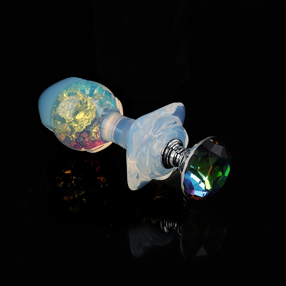 An opal glass butt plug with a colorful rainbow crystal core lying horizontally, featuring a large faceted multicolored rhinestone on the base.