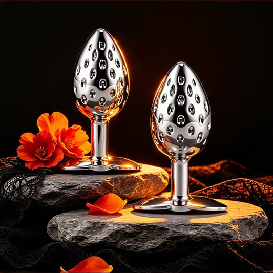 Two strawberry-shaped metal butt plugs with textured surfaces resting on dark stones with orange flowers, showcasing premium body-safe metallic finish.